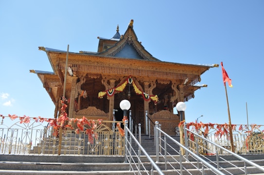 An intricately designed wooden temple featuring ornate carvings and architectural details. The temple is decorated with colorful garlands and is approached by steps with metal railings. A red flag is positioned to the right of the temple, and the surrounding area is adorned with vibrant red and yellow floral decorations.