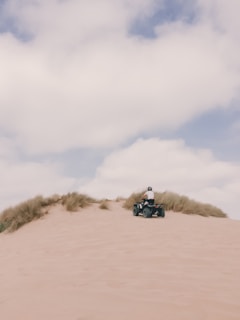 A person riding an ATV on sandy dunes, with tufts of grass and a partly cloudy sky overhead.