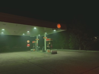 A dimly lit gas station at night with a Shell logo illuminated on the roof. The area appears deserted with no people or vehicles present. A slight fog surrounds the station, casting a green hue. Trees are visible in the background.