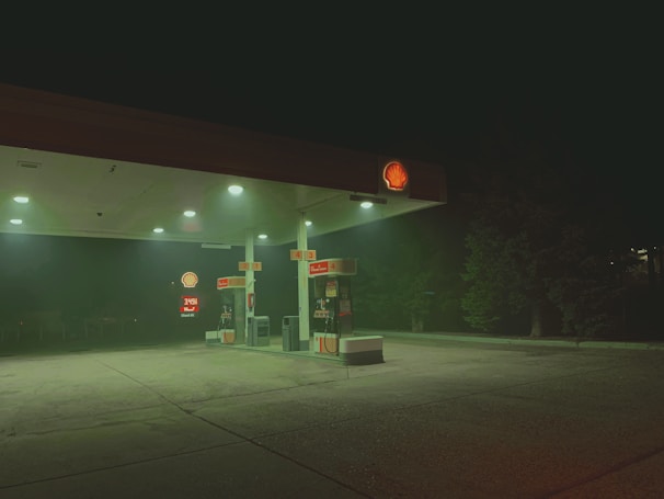 A dimly lit gas station at night with a Shell logo illuminated on the roof. The area appears deserted with no people or vehicles present. A slight fog surrounds the station, casting a green hue. Trees are visible in the background.