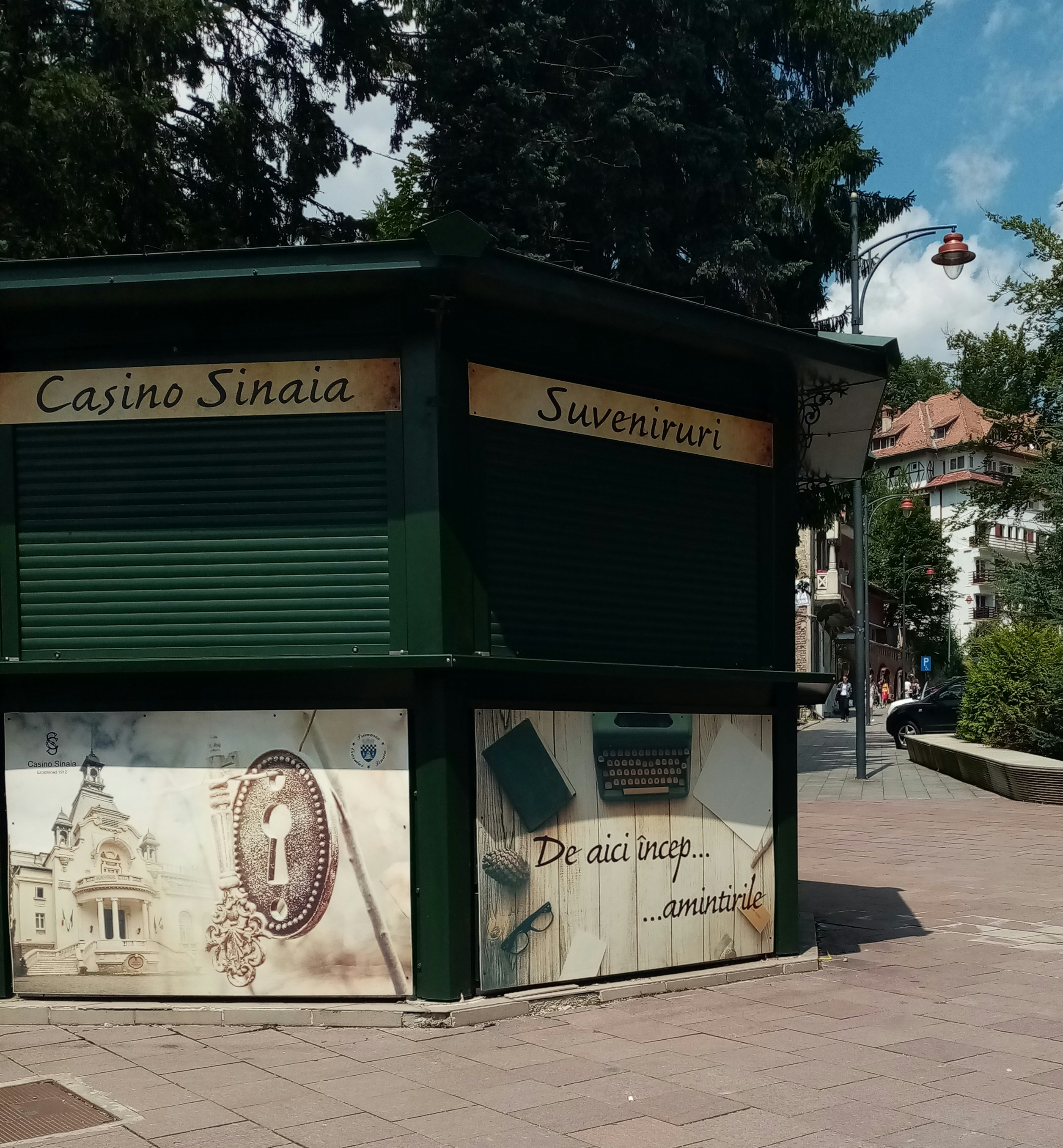 A green kiosk located in an urban outdoor setting, featuring closed rolling shutters. The signage reads 'Casino Sinaia' and 'Suveniruri' indicating it is a souvenir shop. Below the sign, there are decorative panels; one shows an ornate key and building, and another depicts a typewriter and papers with text that translates to 'This is where memories begin.' The scene is surrounded by paved walkways, trees, and partly visible buildings.
