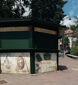 A green kiosk located in an urban outdoor setting, featuring closed rolling shutters. The signage reads 'Casino Sinaia' and 'Suveniruri' indicating it is a souvenir shop. Below the sign, there are decorative panels; one shows an ornate key and building, and another depicts a typewriter and papers with text that translates to 'This is where memories begin.' The scene is surrounded by paved walkways, trees, and partly visible buildings.