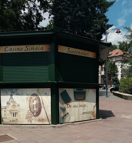 A green kiosk located in an urban outdoor setting, featuring closed rolling shutters. The signage reads 'Casino Sinaia' and 'Suveniruri' indicating it is a souvenir shop. Below the sign, there are decorative panels; one shows an ornate key and building, and another depicts a typewriter and papers with text that translates to 'This is where memories begin.' The scene is surrounded by paved walkways, trees, and partly visible buildings.