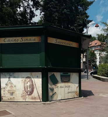 A green kiosk located in an urban outdoor setting, featuring closed rolling shutters. The signage reads 'Casino Sinaia' and 'Suveniruri' indicating it is a souvenir shop. Below the sign, there are decorative panels; one shows an ornate key and building, and another depicts a typewriter and papers with text that translates to 'This is where memories begin.' The scene is surrounded by paved walkways, trees, and partly visible buildings.