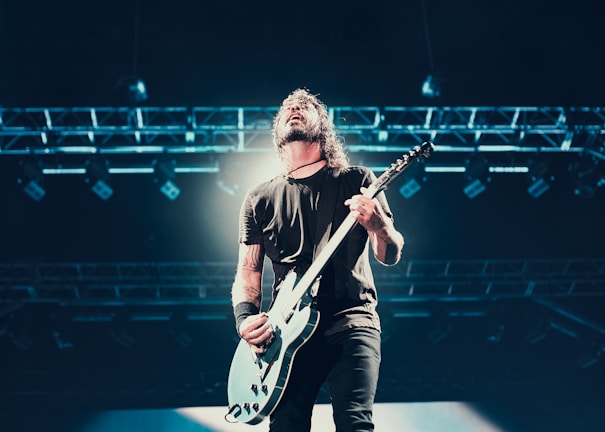 A musician passionately plays an electric guitar on stage under bright lighting. The performer has long hair and is captured mid-performance, with stage lights and rigging visible in the background.