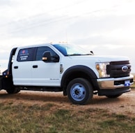 A white utility truck parked on a patch of grass beside a dirt road, set against a lightly clouded sky. The truck features a prominent chrome grille and sturdy tires, with branding and numbers visible on the side.