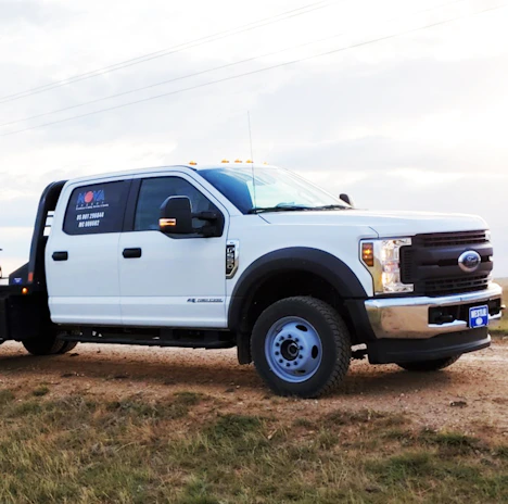 A white utility truck parked on a patch of grass beside a dirt road, set against a lightly clouded sky. The truck features a prominent chrome grille and sturdy tires, with branding and numbers visible on the side.