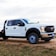 A white utility truck parked on a patch of grass beside a dirt road, set against a lightly clouded sky. The truck features a prominent chrome grille and sturdy tires, with branding and numbers visible on the side.