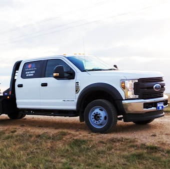 A white utility truck parked on a patch of grass beside a dirt road, set against a lightly clouded sky. The truck features a prominent chrome grille and sturdy tires, with branding and numbers visible on the side.
