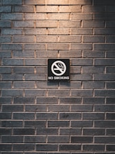 A dark brick wall features a prominently placed 'No Smoking' sign. The sign is centered and displays a cigarette with a red circle and slash, conveying the prohibition.