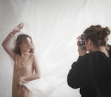 A photographer is capturing a woman posing against a soft, flowing backdrop. The woman is wearing a delicate, sheer dress, and her hair is gently tousled as she strikes an elegant pose. The photographer appears focused while holding a camera up to their eye.