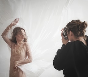 A photographer is capturing a woman posing against a soft, flowing backdrop. The woman is wearing a delicate, sheer dress, and her hair is gently tousled as she strikes an elegant pose. The photographer appears focused while holding a camera up to their eye.