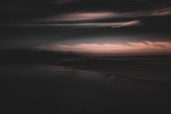 A dark and moody landscape with a dramatic sky filled with soft, swirling clouds. The horizon is bathed in a faint, warm glow, providing a stark contrast to the dark foreground and the expansive cloudy sky.
