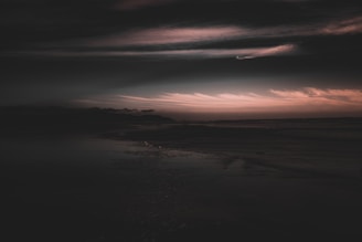 A dark and moody landscape with a dramatic sky filled with soft, swirling clouds. The horizon is bathed in a faint, warm glow, providing a stark contrast to the dark foreground and the expansive cloudy sky.