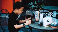 A young boy is seated at an outdoor cafe table, intently engaged with a tablet. The table holds a glass of iced coffee with a straw, a menu, a tissue holder, and a cupcake. The surroundings feature vintage-style wrought iron chairs with some plants in the background.
