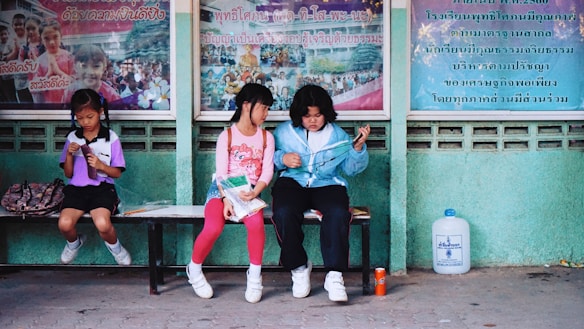 Three children sit on a bench, engaged in different activities. The child on the left, wearing a purple shirt, is reading a book. The child in the center, wearing a pink shirt, is holding papers and looking at the child on the right, who wears a light blue jacket and appears to be tying a string. Behind them, there are colorful posters with text and images. A large water container and a small orange can are on the ground nearby.