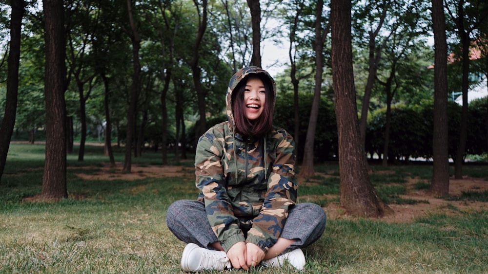 Femme en sweat à capuche de camouflage et pantalon gris assis sur des herbes près des arbres