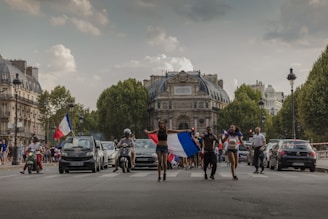 A group of people celebrating in the middle of a street, some holding French flags. Cars and scooters are nearby, and a historic building stands in the background, lined with large trees on each side of the street.