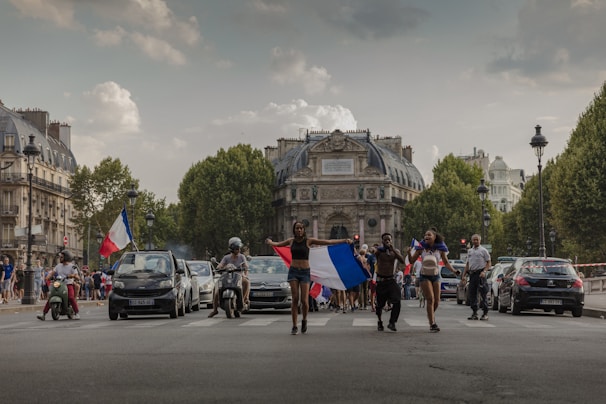 A group of people celebrating in the middle of a street, some holding French flags. Cars and scooters are nearby, and a historic building stands in the background, lined with large trees on each side of the street.
