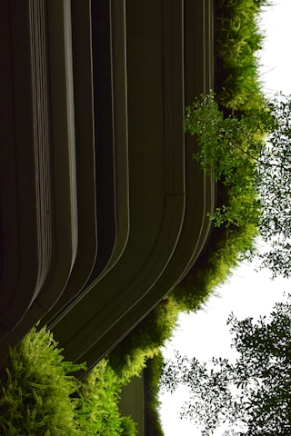 Curved architectural structures are integrated with lush greenery, appearing as a blend between modern design and natural elements. The greenery comprises various shades of vibrant green plants cascading down from the building.