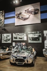 A vintage race car is prominently displayed in a museum setting. The glossy vehicle sits on a polished concrete floor, surrounded by framed black and white photographs of similar racing cars. Above it, a large sepia-toned image of two classic race cars hangs on a dark wall.