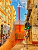 A hand holds a cup of bubble tea in front of a vibrant urban street scene, with tall, colorful buildings lining the street. A red straw sticks out from the cup, and there is a traffic light showing green for pedestrians in the background. Several people can be seen walking along the sidewalk.