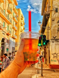 A hand holds a cup of bubble tea in front of a vibrant urban street scene, with tall, colorful buildings lining the street. A red straw sticks out from the cup, and there is a traffic light showing green for pedestrians in the background. Several people can be seen walking along the sidewalk.
