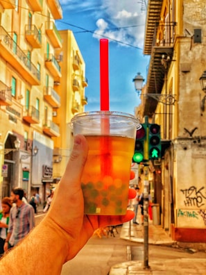 A hand holds a cup of bubble tea in front of a vibrant urban street scene, with tall, colorful buildings lining the street. A red straw sticks out from the cup, and there is a traffic light showing green for pedestrians in the background. Several people can be seen walking along the sidewalk.