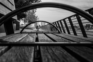 A black and white perspective shot looking through a metal circular structure on a wooden bench. The image captures the lines and texture of the wood and the metal, with a blurred background including railings, a pathway, and trees. The scene conveys depth and symmetry.