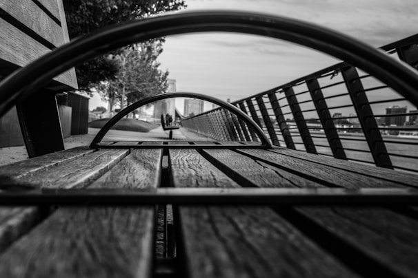 A black and white perspective shot looking through a metal circular structure on a wooden bench. The image captures the lines and texture of the wood and the metal, with a blurred background including railings, a pathway, and trees. The scene conveys depth and symmetry.