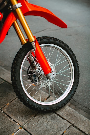 A close-up view of a motorcycle wheel with a knobby tire parked on a stone pavement. The tire is clean, and the spokes are shiny, reflecting light. The red and gold components of the motorcycle frame are visible, indicating a sporty and vibrant aesthetic.