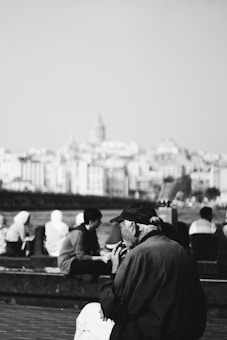An elderly man sits by the waterfront, looking contemplative. The background features a cityscape with a prominent tower, and several other people are sitting or walking nearby, some with their backs to the camera.