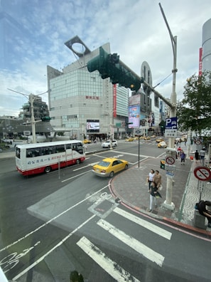 A bustling urban intersection with a large modern building in the background featuring digital billboards. Various vehicles, including a red and white bus and a yellow taxi, are on the road. Pedestrians are crossing the street, and traffic signals are visible.
