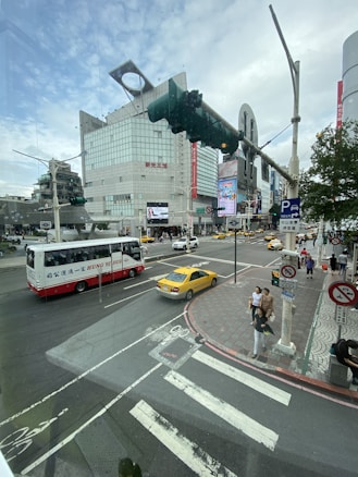 A bustling urban intersection with a large modern building in the background featuring digital billboards. Various vehicles, including a red and white bus and a yellow taxi, are on the road. Pedestrians are crossing the street, and traffic signals are visible.