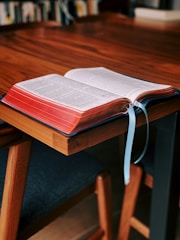 An open book rests on a wooden table, with its pages visible and two blue ribbons marking places. The book appears thick with red edges, and the setting seems to be a study or library, indicated by blurred bookshelves in the background.