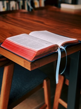 An open book rests on a wooden table, with its pages visible and two blue ribbons marking places. The book appears thick with red edges, and the setting seems to be a study or library, indicated by blurred bookshelves in the background.