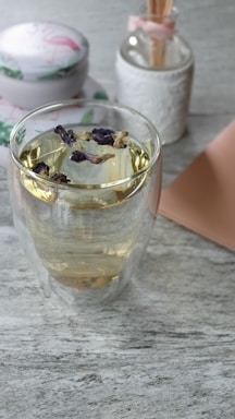 A clear glass cup filled with light yellow tea adorned with dried flowers sits on a gray marble surface. In the background, a decorative jar with a pink flamingo design and a white vase with a pink ribbon border complete the serene setup.