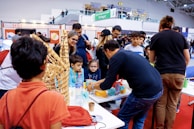 A group of people gather around a table showcasing crafts or educational projects. Children and adults are engaged in observing and interacting with the presented items. The setting appears to be a lively indoor event, possibly a fair or exhibition.