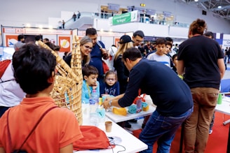 A group of people gather around a table showcasing crafts or educational projects. Children and adults are engaged in observing and interacting with the presented items. The setting appears to be a lively indoor event, possibly a fair or exhibition.