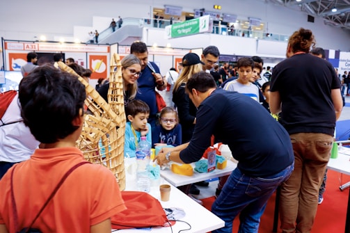 A group of people gather around a table showcasing crafts or educational projects. Children and adults are engaged in observing and interacting with the presented items. The setting appears to be a lively indoor event, possibly a fair or exhibition.