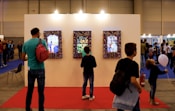 A group of people stand in front of a large exhibition wall featuring three framed digital displays with vibrant artwork. The exhibition is set up indoors with a blue carpet and people are scattered across the background, indicating a busy event or gathering. The lighting is warm, and there are various individuals of different ages observing the displays.
