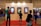 A group of people stand in front of a large exhibition wall featuring three framed digital displays with vibrant artwork. The exhibition is set up indoors with a blue carpet and people are scattered across the background, indicating a busy event or gathering. The lighting is warm, and there are various individuals of different ages observing the displays.
