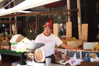 A vendor is sitting at a market stall surrounded by cardboard boxes filled with pineapples and other fruits. He is wearing a white T-shirt and a red and black cap. There is an old-fashioned weighing scale on the table in front of him.