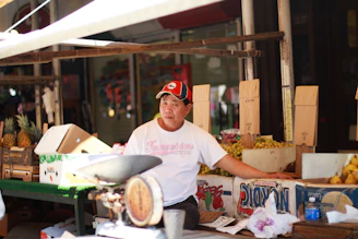 A vendor is sitting at a market stall surrounded by cardboard boxes filled with pineapples and other fruits. He is wearing a white T-shirt and a red and black cap. There is an old-fashioned weighing scale on the table in front of him.