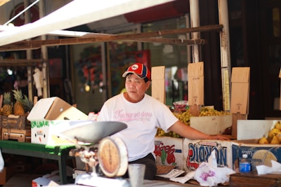 A vendor is sitting at a market stall surrounded by cardboard boxes filled with pineapples and other fruits. He is wearing a white T-shirt and a red and black cap. There is an old-fashioned weighing scale on the table in front of him.