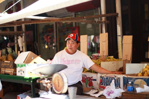 A vendor is sitting at a market stall surrounded by cardboard boxes filled with pineapples and other fruits. He is wearing a white T-shirt and a red and black cap. There is an old-fashioned weighing scale on the table in front of him.