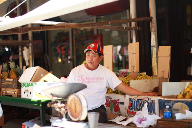 A vendor is sitting at a market stall surrounded by cardboard boxes filled with pineapples and other fruits. He is wearing a white T-shirt and a red and black cap. There is an old-fashioned weighing scale on the table in front of him.