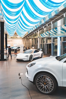 A modern showroom features two sleek white sports cars displayed under an elegant ceiling with blue and white striped fabric. The polished floor reflects the ambient light, and a railing separating the first and second floors adds a touch of architectural sophistication. A person stands near one of the cars, possibly an employee or customer, emphasizing the environment's professional and luxurious feel.