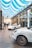 A modern showroom features two sleek white sports cars displayed under an elegant ceiling with blue and white striped fabric. The polished floor reflects the ambient light, and a railing separating the first and second floors adds a touch of architectural sophistication. A person stands near one of the cars, possibly an employee or customer, emphasizing the environment's professional and luxurious feel.
