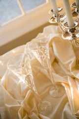 Two crystal champagne flutes are placed on an elegant cream-colored fabric near a silver candle holder. The afternoon sunlight casts soft reflections and highlights, contributing to a warm and sophisticated ambiance.